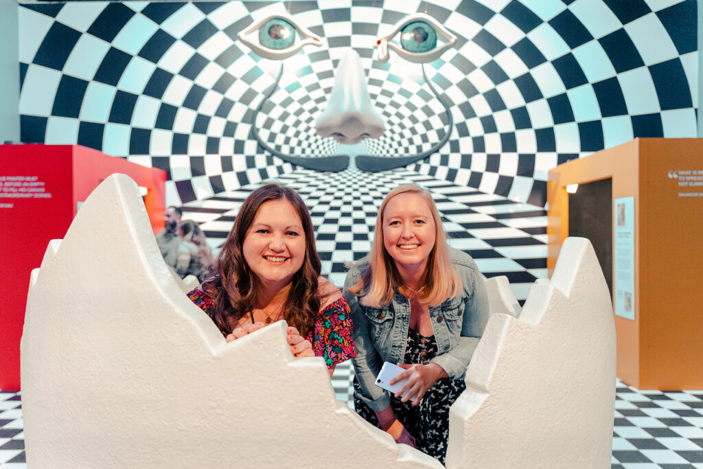 Two people sat inside the Dali Alive Egg with a checkerboard background and Salvador Dali face image by Grande Experiences